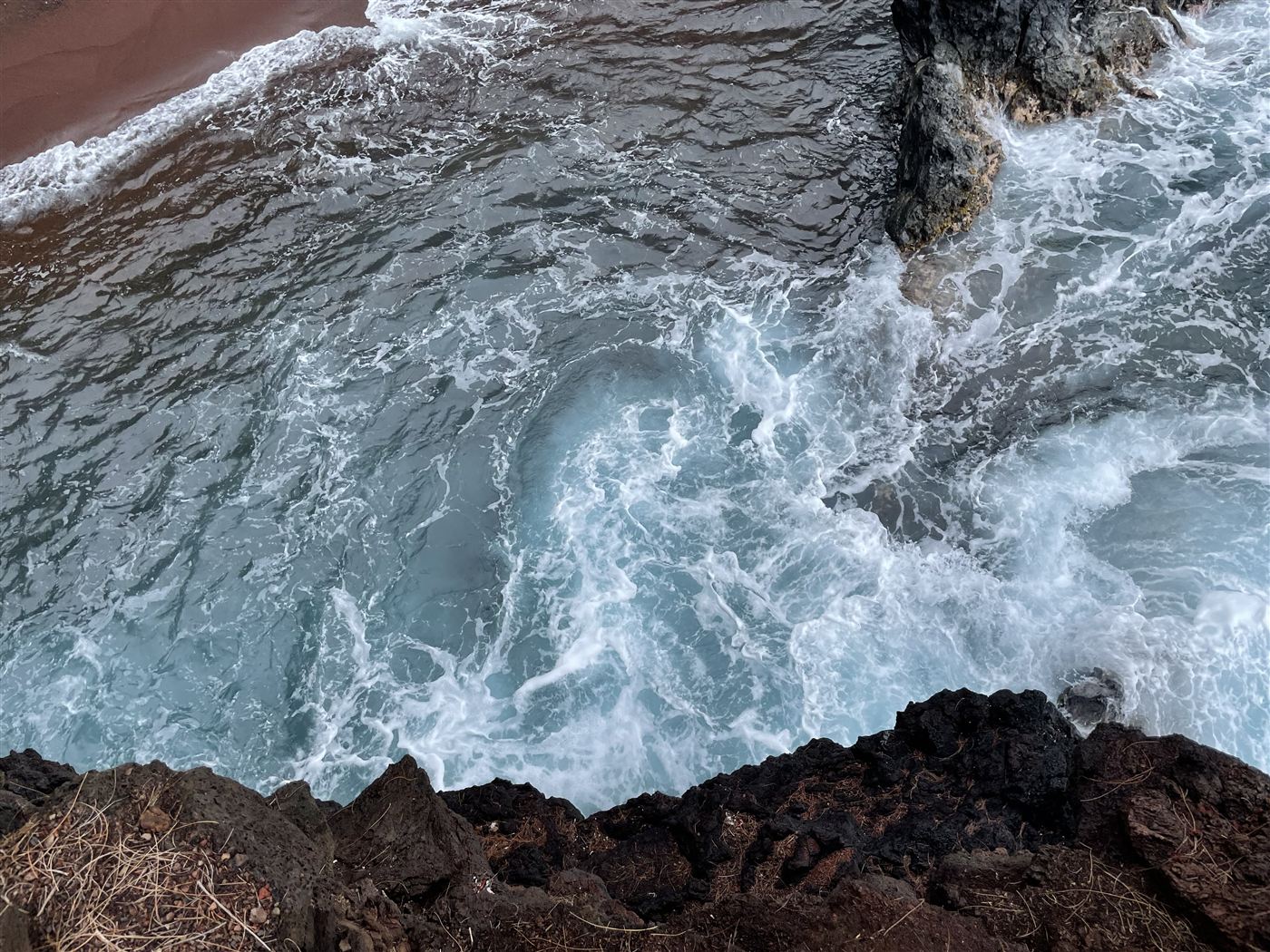 Surf foaming against volcanic rock above a red sand beach, seen from above